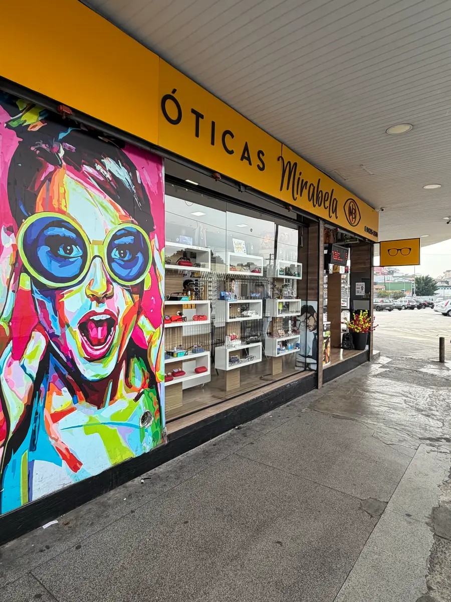 Fachada da unidade Vila Luzita das Óticas Mirabela, com letreiro amarelo, vitrine de armações e mural colorido na lateral